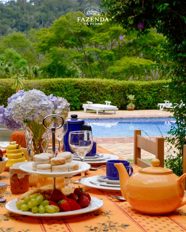 Mesa de café da manhã com hortênsias, bule laranja e frutas, de frente para a piscina