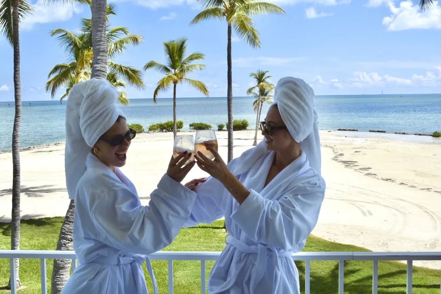 Duas mulheres de roupão brindando na varanda, com a praia e os coqueiros ao fundo